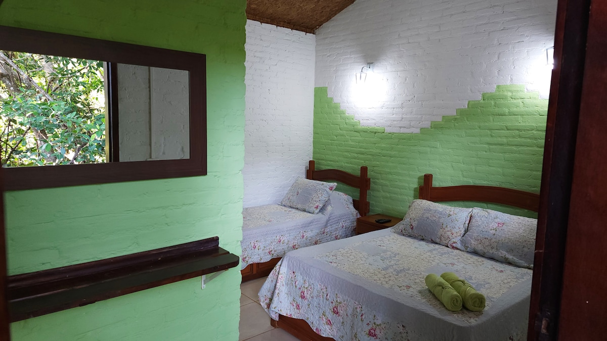 A cozy bedroom is presented, featuring a double bed and a single bed, both adorned with floral bed linens. A mirror is mounted on a green wall, and light fixtures illuminate the space. Fresh towels are neatly arranged on the beds, enhancing the relaxed ambiance.