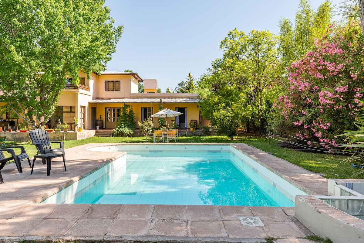 A tranquil garden scene is captured, featuring a spacious swimming pool surrounded by stone pathways. Lush greenery and colorful flowers provide a natural backdrop, with comfortable seating visible to one side. The house stands proudly in the background, integrating with the serene outdoor space.