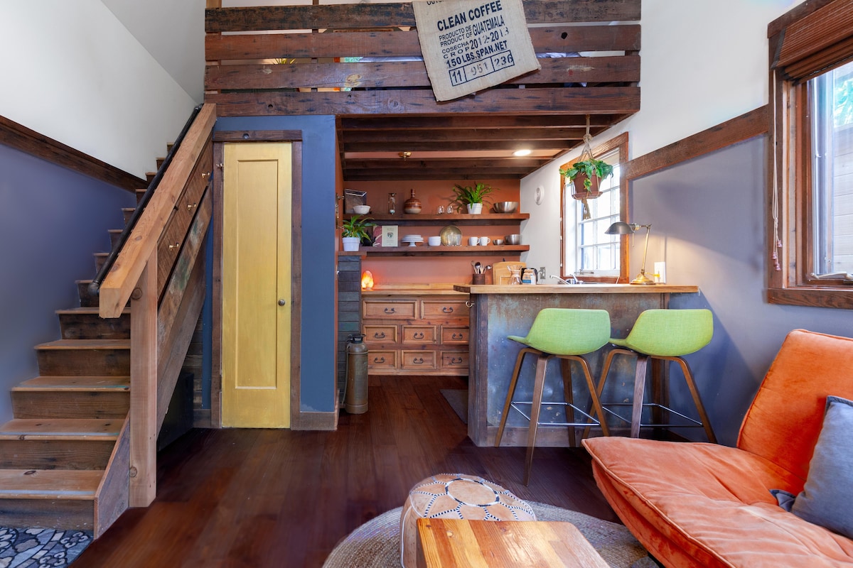 The interior features a rustic aesthetic with wooden staircase and walls. A compact kitchen area showcases a bar with two green stools, surrounded by various plants. An inviting orange futon is positioned in the living space, complemented by a wooden coffee table.