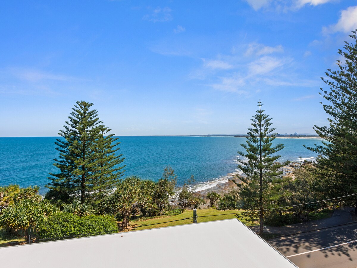 A panoramic view of the ocean is visible, framed by tall palm trees and lush greenery. The water glistens under the sunlight, and the shoreline can be seen merging with the gentle waves. A clear blue sky enhances the tranquil coastal scene.