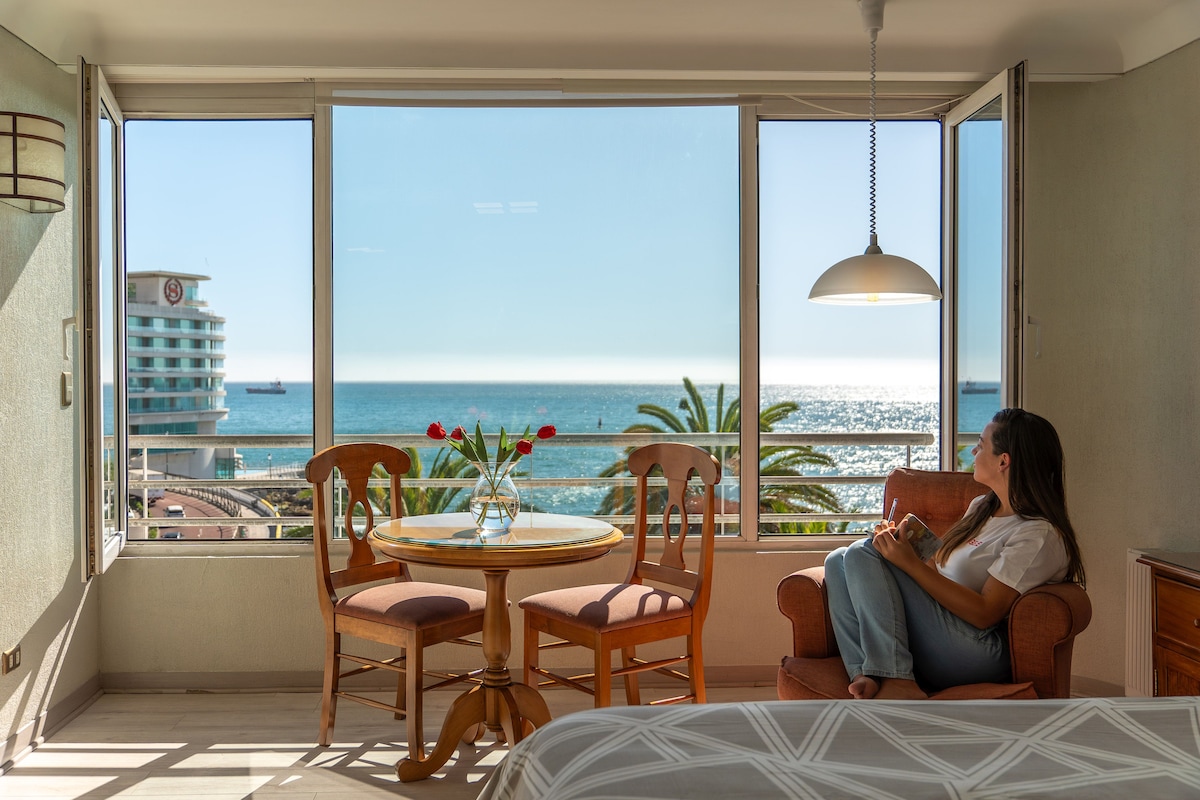 A bright studio apartment is showcased with large windows offering a scenic view of the ocean. A round dining table with two chairs is positioned near the window, complemented by a comfortable armchair. Natural light fills the space, highlighting the coastal scenery.