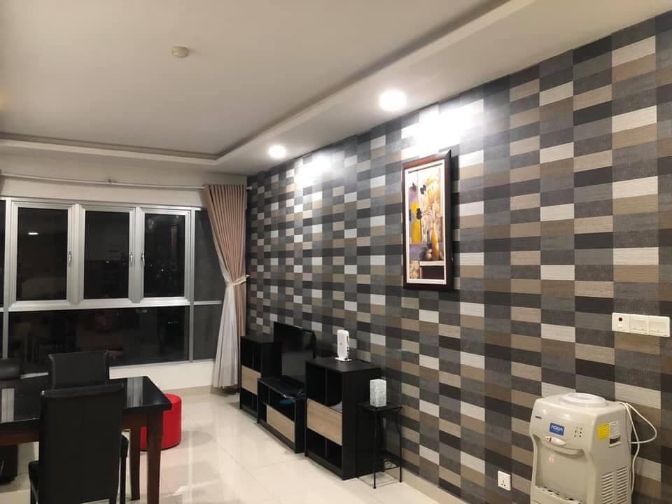 A modern living space features a patterned accent wall in neutral tones, framed artwork, and a window with natural light. A dining area with a table and chairs is positioned beside a compact shelving unit, providing organization and style. Air conditioning is seen in the corner.