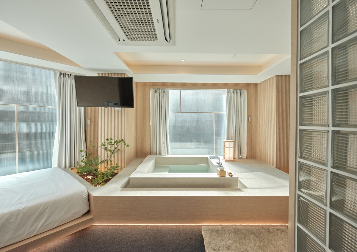 A spacious studio room is highlighted by natural light filtering through large windows. A cozy sleeping area features a bed with a soft white cover. A jacuzzi is integrated into the design, surrounded by minimalist decor and a small indoor plant, creating a serene environment.