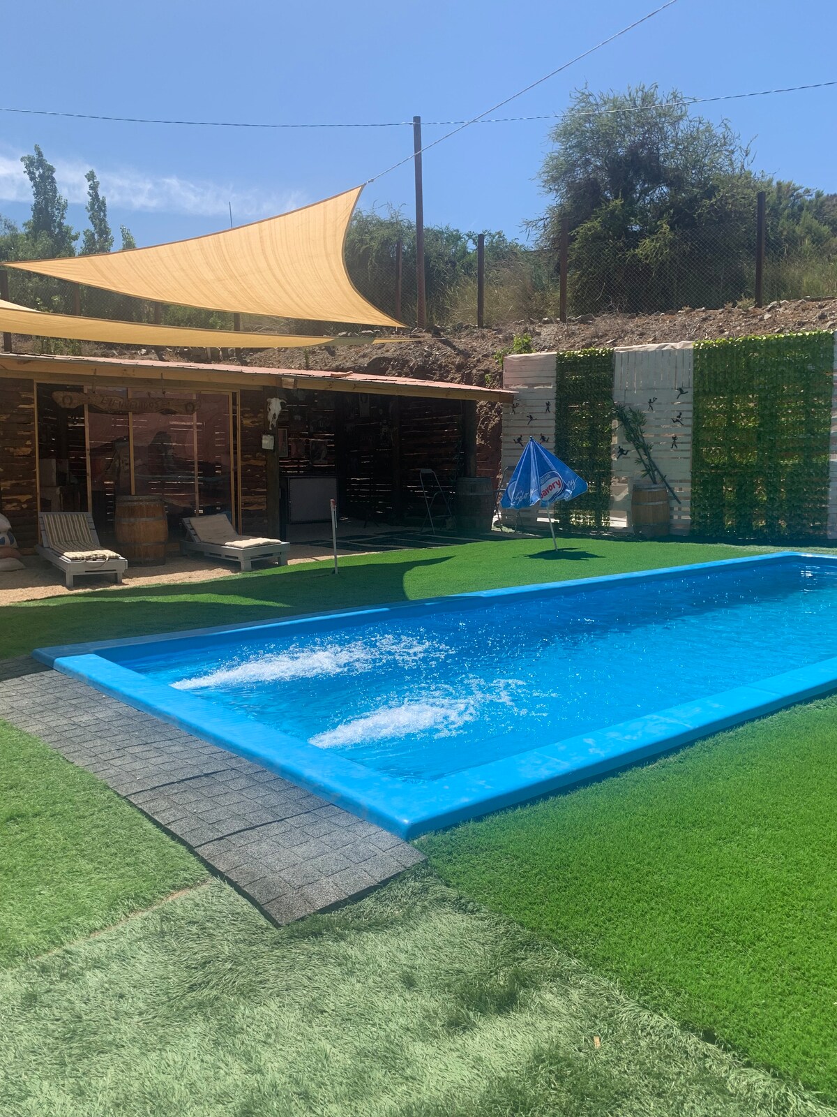 A refreshing pool glistens under the sunlight, surrounded by artificial grass and shaded by a large canopy. Lounge chairs are positioned nearby, providing a serene space for relaxation. The area is complemented by natural stone elements in the background.