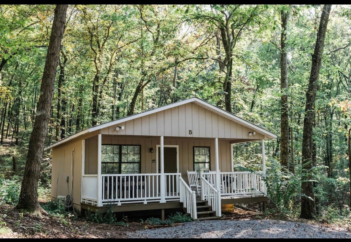 A cabin is set against a backdrop of lush trees, featuring a covered porch with white railings. Large windows allow natural light to fill the interior. A gravel driveway enhances the inviting entrance to this serene woodland retreat.