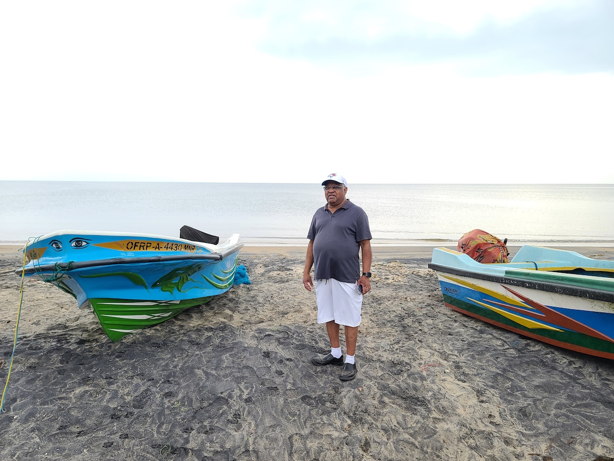 Two vibrant fishing boats are positioned on a sandy beach, with water gently rippling in the background. A man stands between the boats, dressed in casual attire, as soft clouds float above in an expansive sky.