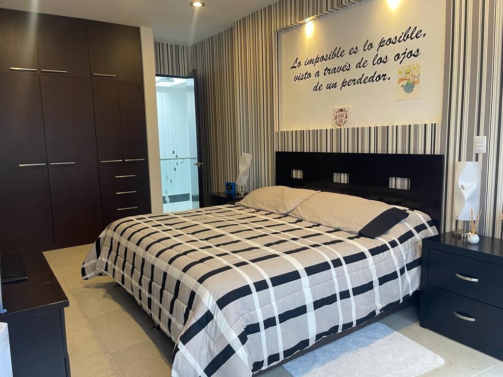 A bedroom is presented with a spacious bed, dressed in a striped black and white blanket. The room features patterned wallpaper in neutral tones and built-in wardrobes. Bedside tables with modern lamps are positioned on either side of the bed, contributing to a functional layout.