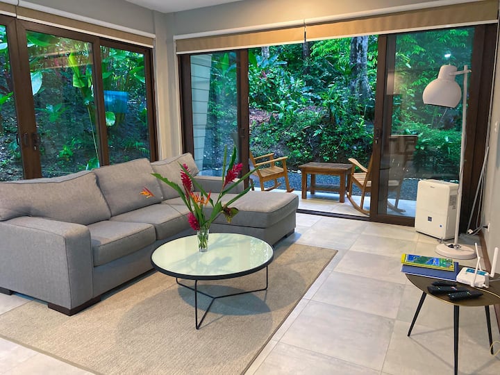 La photo montre un salon costaricien moderne. La chambre est lumineuse et aérée avec de grandes fenêtres donnant sur une végétation luxuriante. Un canapé sectionnel confortable, une table basse ronde et une table d'appoint avec une lampe complètent la configuration confortable.