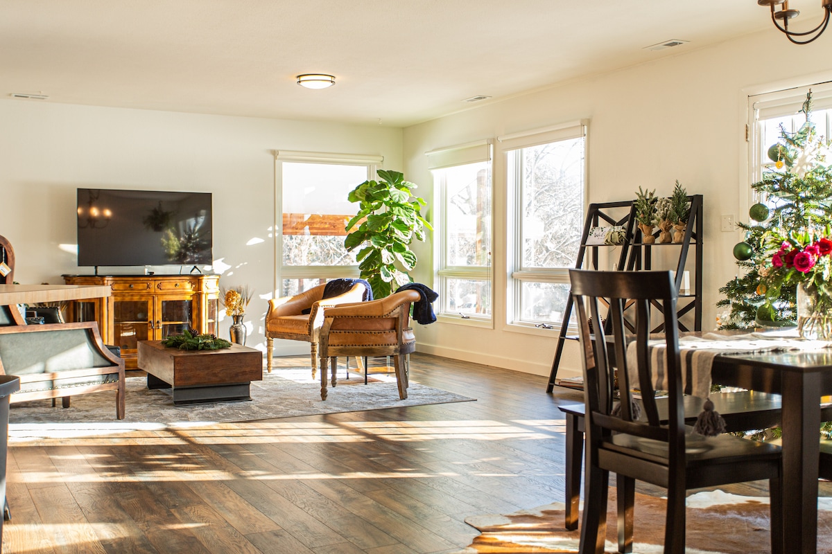 A bright living area features large windows allowing natural light to fill the space. Comfortable seating is provided by two chairs alongside a wooden table. A television is mounted on the wall, while a small dining table sits nearby, adorned with a centerpiece of flowers.