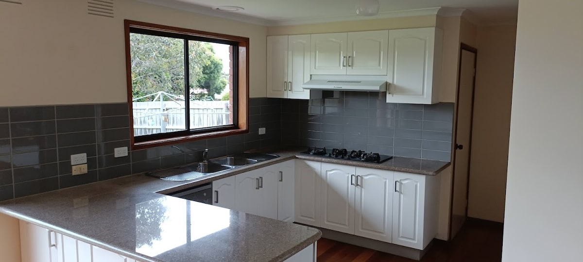 A modern kitchen features white cabinetry and a spacious granite countertop. The kitchen is equipped with stainless steel appliances, including a gas stove. A large window allows natural light to illuminate the room, with a view of greenery outside.
