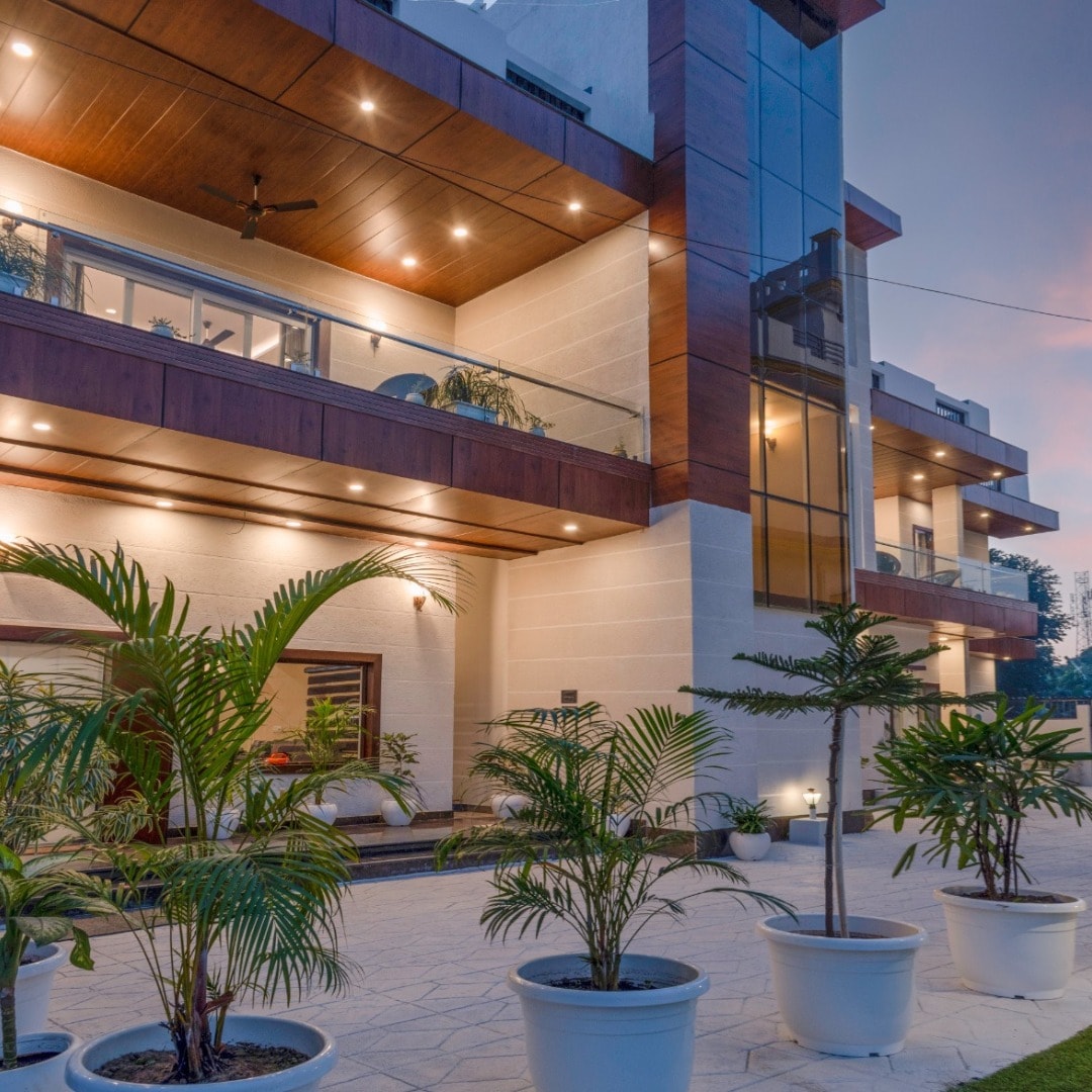 The exterior of the villa features a modern design with a combination of concrete and wood accents. Lush green plants in white pots frame the entrance, and large glass windows reflect the soft evening light, enhancing the welcoming ambiance of the property.