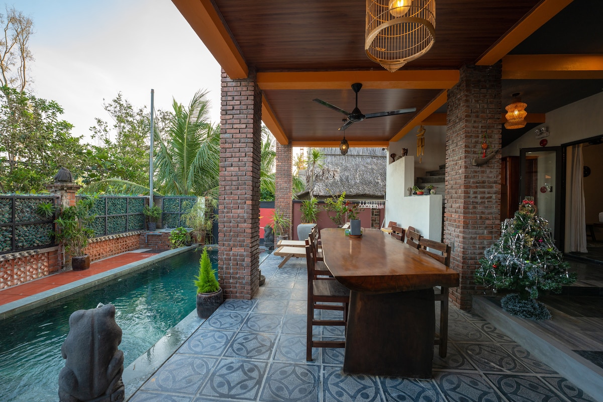 A covered outdoor dining area features a large wooden table surrounded by several chairs, set alongside a private swimming pool. Lush greenery is visible in the background, and decorative lighting adds ambiance to the space.