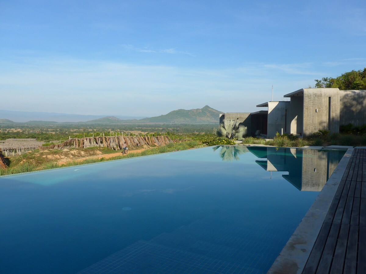 An infinity swimming pool extends towards the horizon, reflecting clear blue skies. Surrounding mountainous landscapes and greenery are visible in the background, enhancing the serene setting. The modern architecture of a nearby building complements the natural beauty.