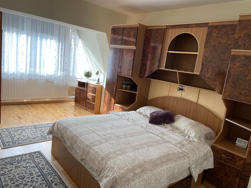 A well-lit bedroom features a spacious bed with neutral linens and a decorative pillow. Wood cabinetry lines the wall, providing storage, while a desk and chair are positioned nearby. Natural light filters through sheer curtains, adding a bright touch to the inviting space.