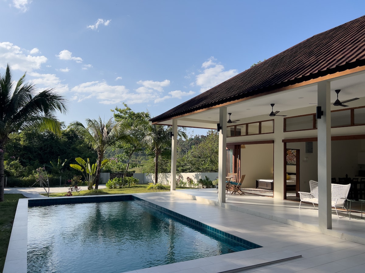A private pool with a shimmering surface is framed by lush greenery and a modern villa. The villa features a broad overhanging roof and sliding doors that open to the outdoor area, allowing for plenty of natural light. A serene atmosphere is created by surrounding palms and calming mountain views.