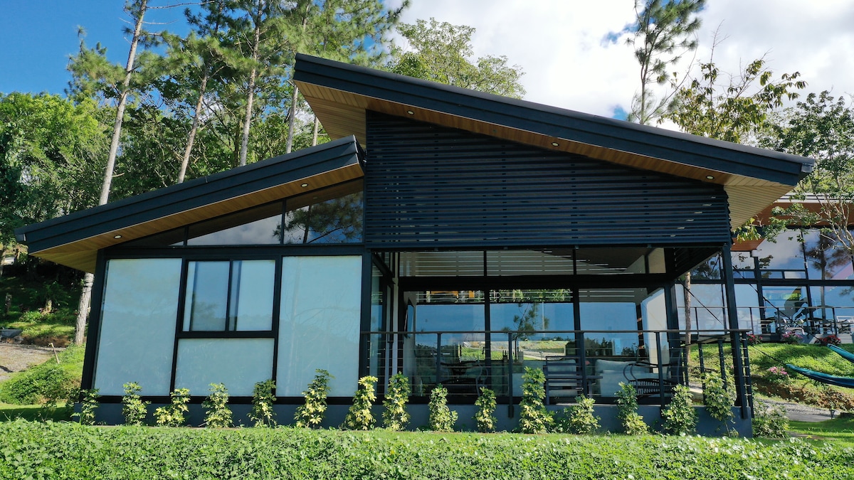 A modern cabin is set against a backdrop of lush greenery, featuring a sleek exterior design with large glass windows. A spacious terrace is visible, lined with vibrant plants, while the angled roof adds visual interest. The serene landscape complements the structure's contemporary style.