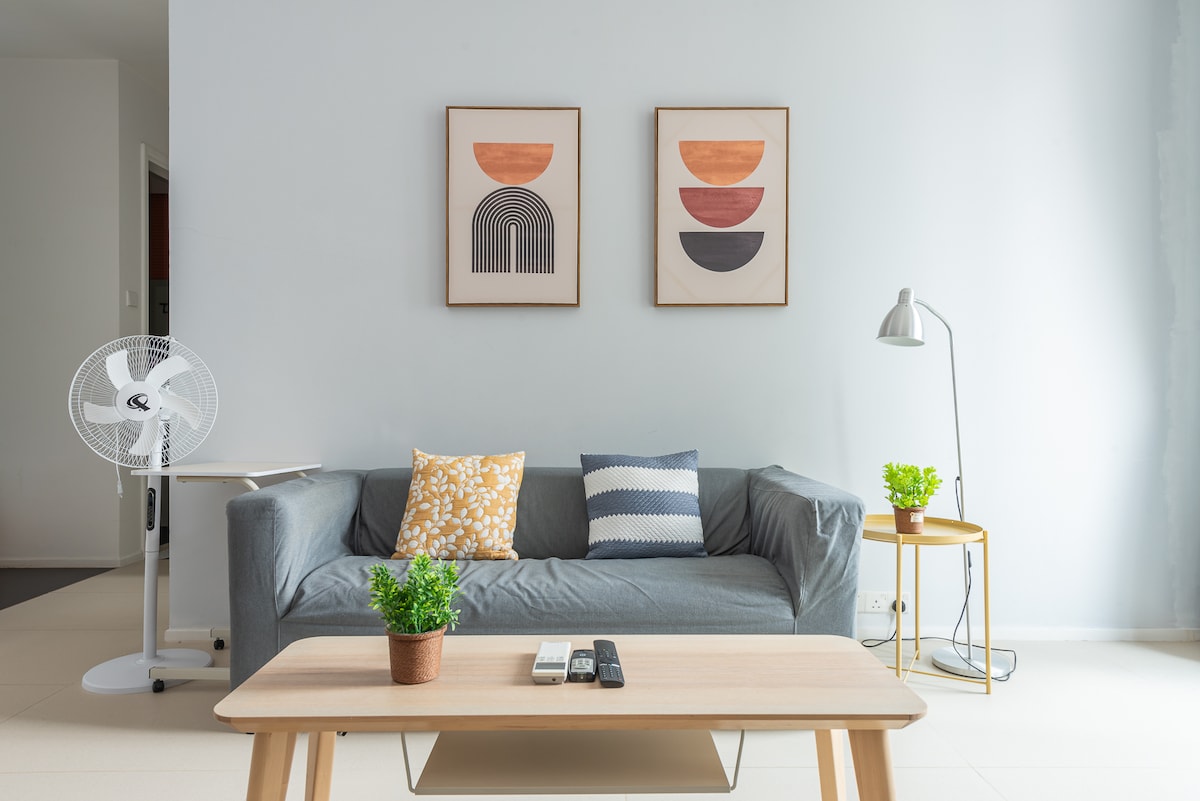 A modern living area features a comfortable gray sofa adorned with colorful cushions. Above the sofa, two framed art pieces are displayed on the pale wall. A wooden coffee table is positioned in front, complemented by a small potted plant and remote controls. A standing lamp and a fan are also visible.