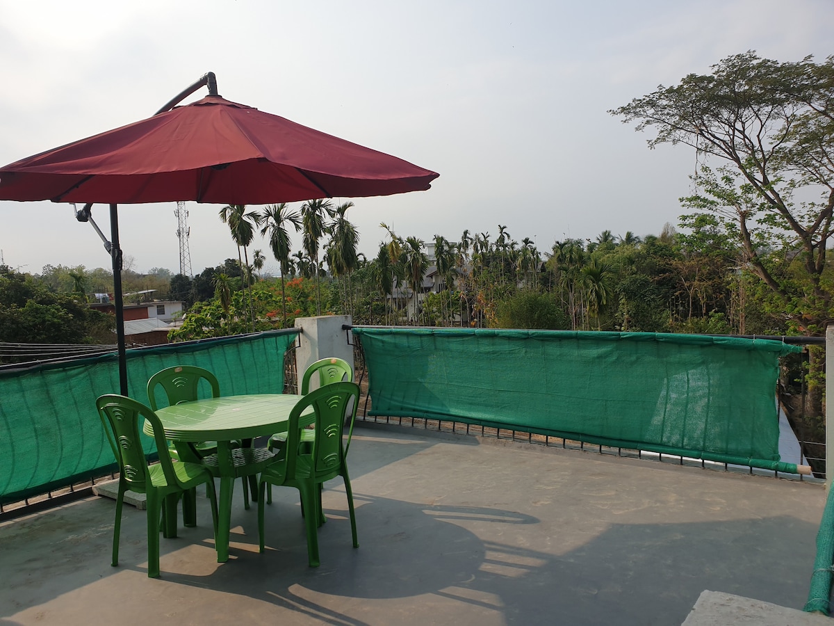 A rooftop terrace features a round table surrounded by four green chairs, shaded by a large red umbrella. Green mesh fencing provides a sense of privacy while offering views of surrounding trees and rooftops.