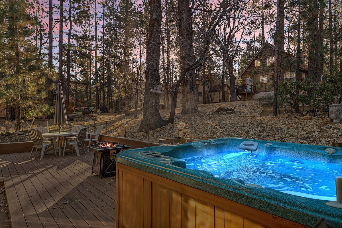 A wooden deck features a hot tub with inviting blue water, surrounded by tall trees. A fire pit is visible nearby, along with chairs and a table, creating an outdoor space suitable for relaxation and gatherings.