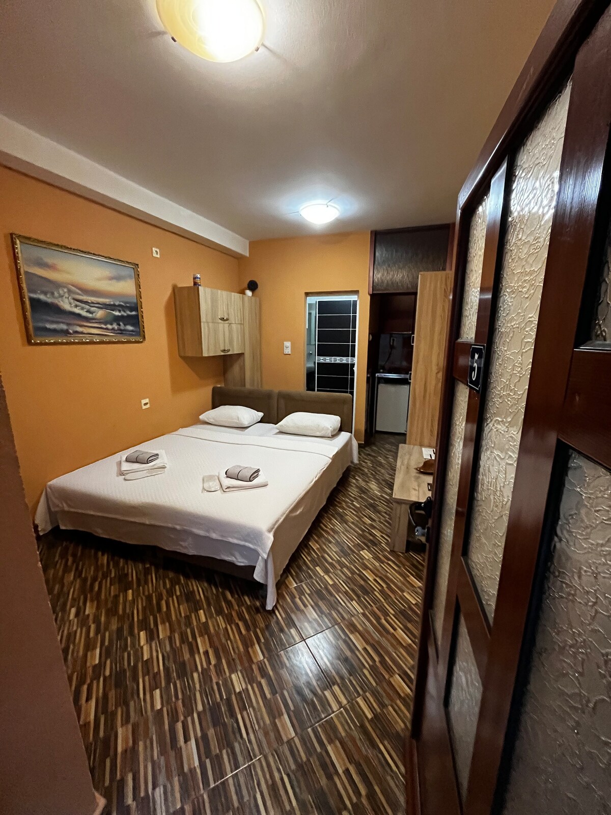 A cozy sleeping area is furnished with a spacious bed covered in a white linen set. Natural wood accents are present in the cabinetry and flooring, complemented by warm orange walls. Soft lighting illuminates the room, highlighting a small window and decorative artwork.