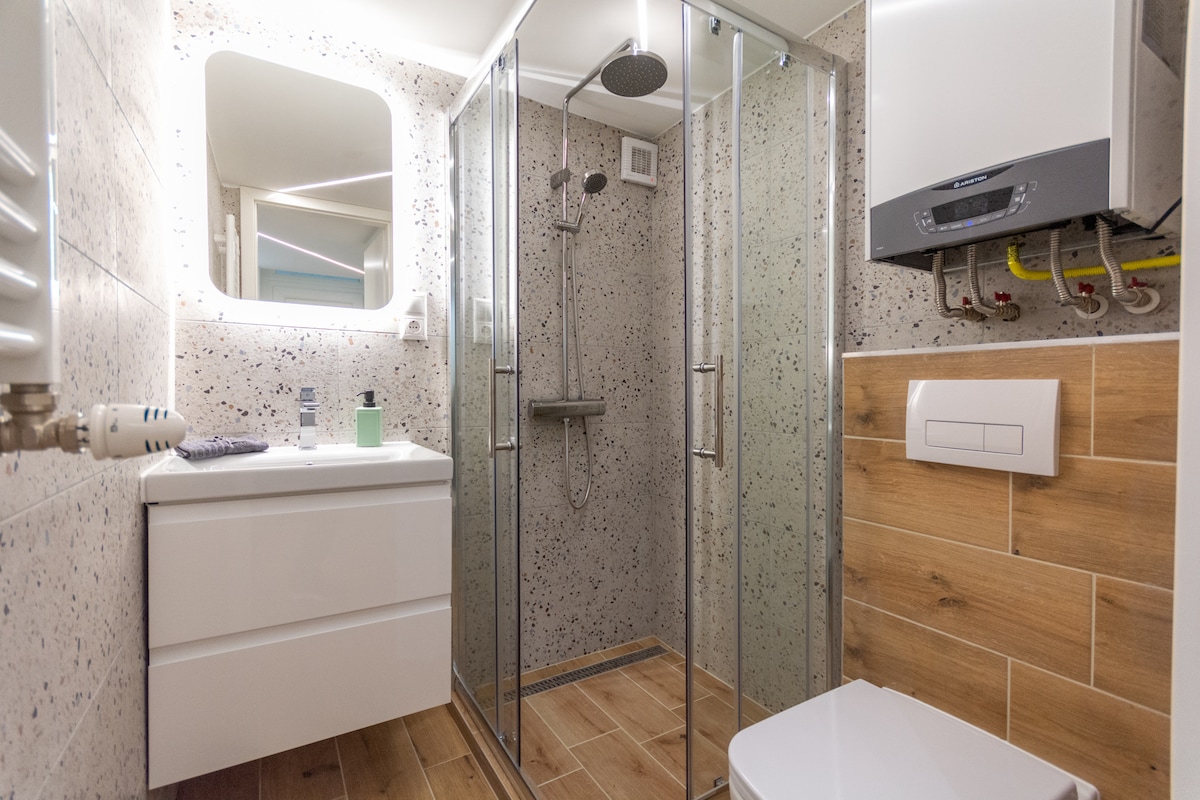 A modern bathroom is featured, showcasing a spacious glass shower with a rainfall showerhead. A sleek white vanity with a mirror is present, alongside a compact toilet. Stylish tile work in neutral tones enhances the clean, contemporary design.