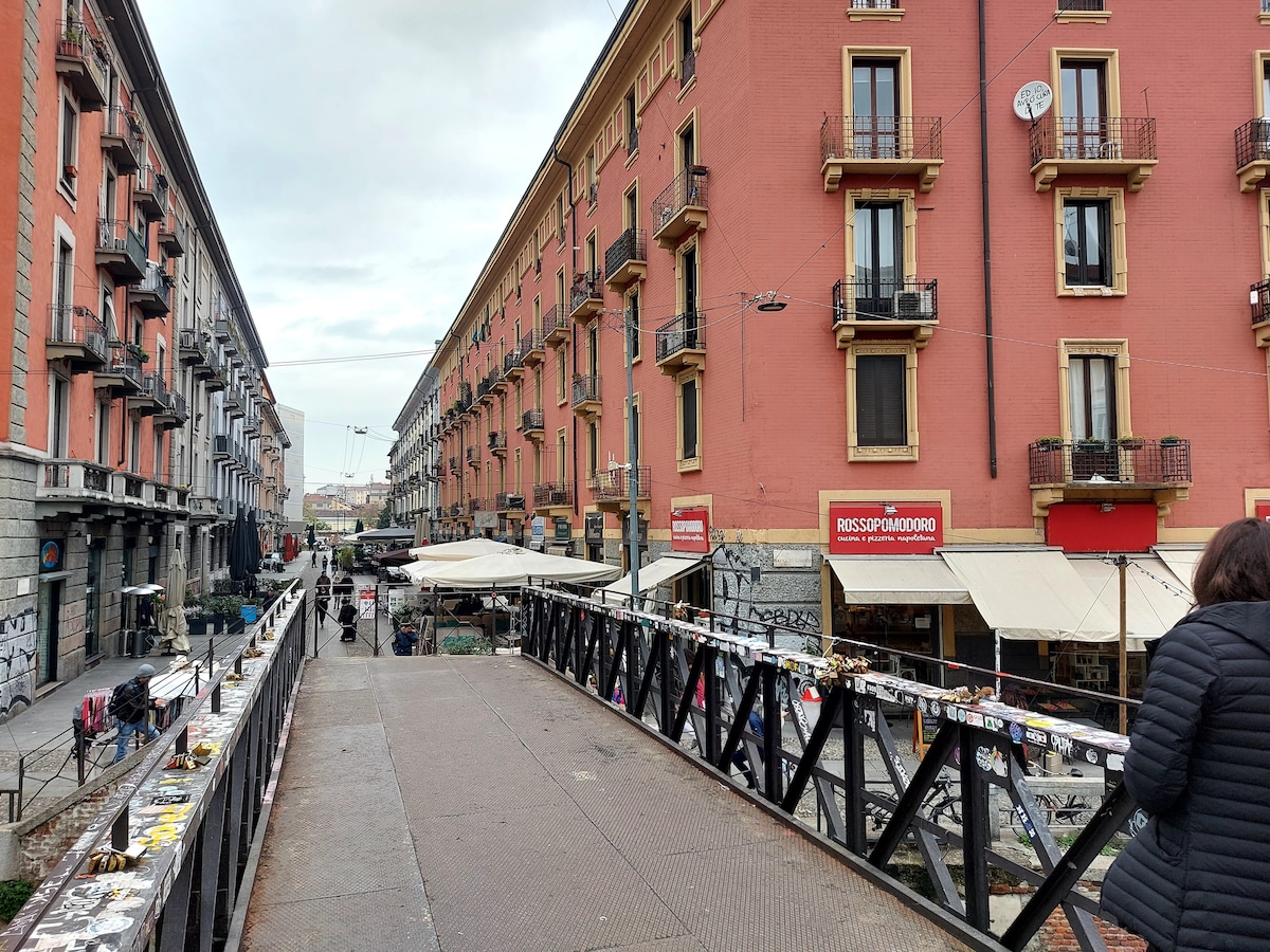 A view of a charming street lined with buildings featuring various balcony styles and colorful facades. A bridge connects the street, with outdoor seating visible at cafes below. None of the streets are crowded, reflecting a calm atmosphere in this vibrant area.