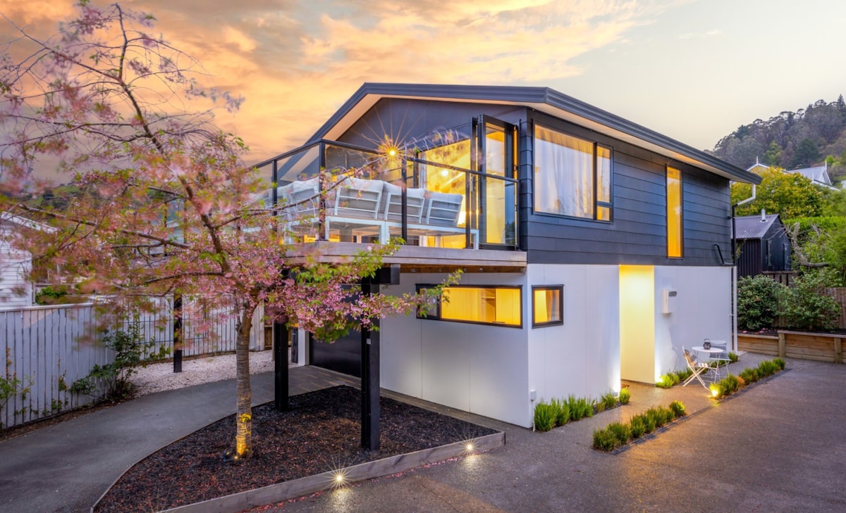 A modern two-story home is featured, with a wraparound balcony surrounded by glass railing. A cherry blossom tree sits in front, complementing the soft lighting from the building. The clean lines and contrasting colors of the exterior create an appealing contemporary aesthetic.
