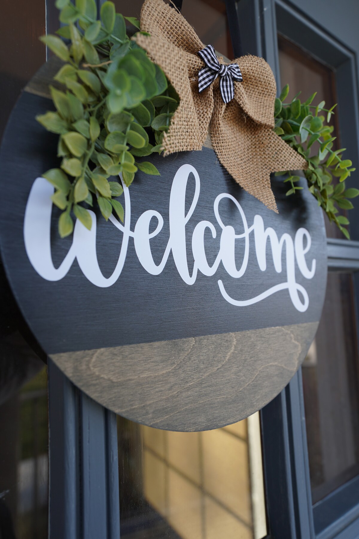 A welcome sign is displayed on the door, featuring a black background with white lettering. The sign is adorned with greenery and a burlap bow, adding a touch of warmth to the entrance.