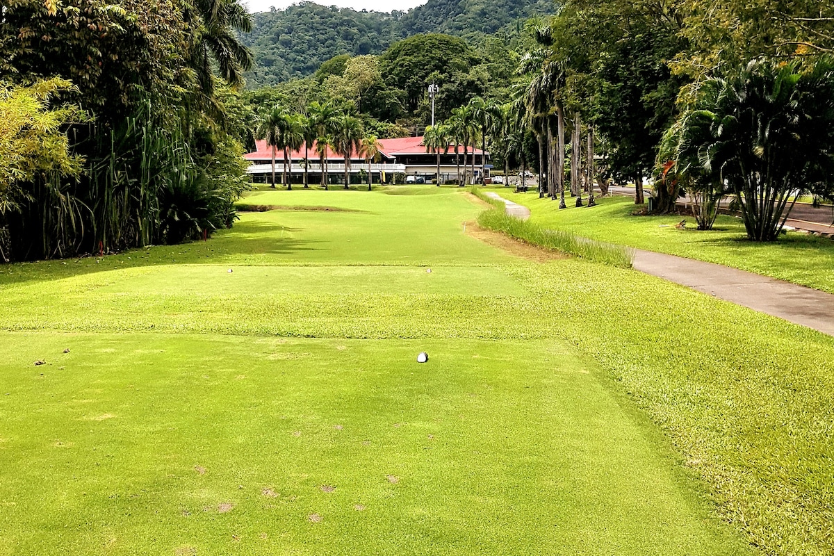 A well-maintained golf course is depicted, featuring lush green fairways bordered by palm trees and tropical foliage. A clubhouse is visible in the background, complemented by a pathway leading to it. The scenery is surrounded by verdant hills, creating a serene golfing environment.