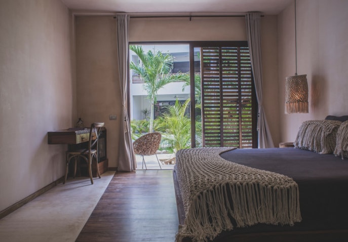 A spacious bedroom is illuminated by natural light from a large glass door opening to a private terrace. Features a large bed adorned with textured textiles, a cozy sitting area with woven chairs, and a wooden desk against the wall. Lush greenery can be seen outside.