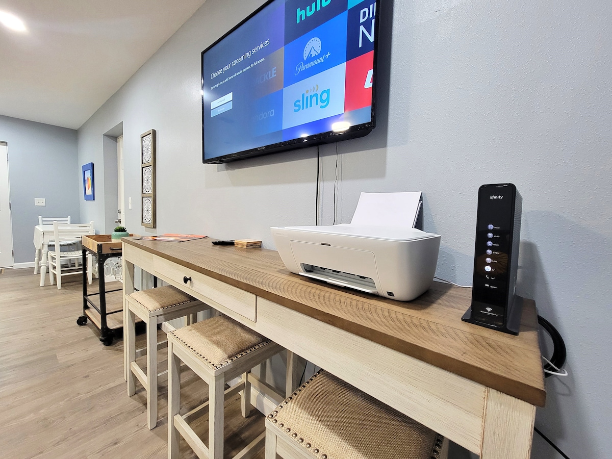 A modern workspace is featured with a wooden table and two stools. A printer sits beside a digital device on the right. A large television is mounted on the wall, displaying various streaming service icons, enhancing the functional atmosphere.