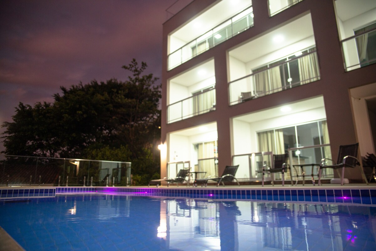 A modern building is reflected in the tranquil water of an infinity pool, surrounded by lounge chairs. The structure features multiple balconies with glass railings, illuminated by soft lighting, and is set against a backdrop of greenery under a twilight sky.
