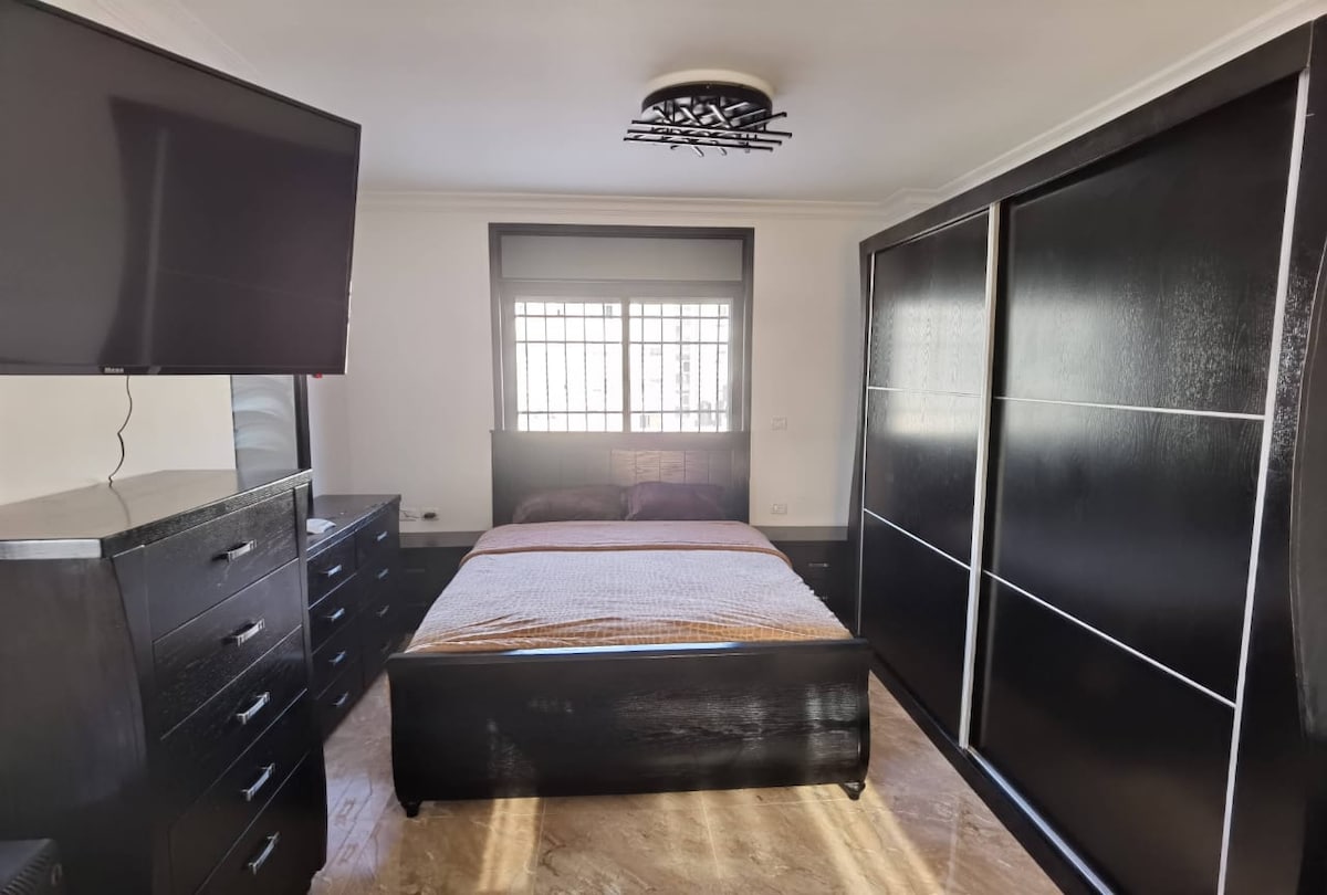 A well-appointed bedroom is equipped with a spacious bed covered in a neutral-toned blanket. A large flat-screen television is mounted on the wall opposite. Built-in wardrobes provide ample storage, while natural light filters through the window, framed by a grid pattern.