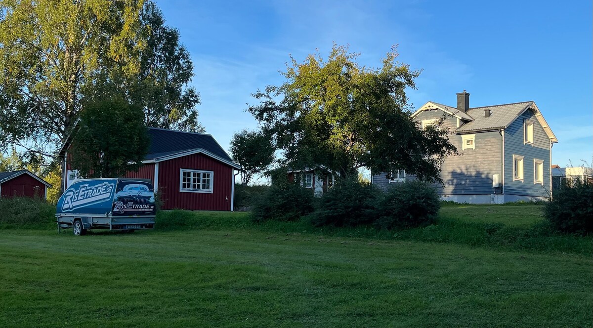 A green lawn stretches across the foreground, with a view of two buildings: a red barn and a gray house. A parked vehicle is visible next to the barn, with clear skies and trees framing the scene.