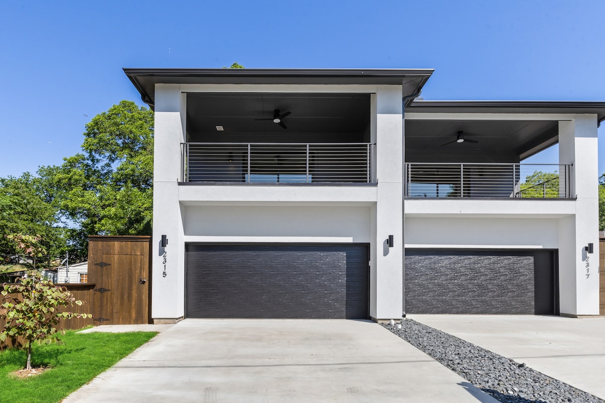 The modern exterior of the property features two spacious garages with sleek black doors. A covered balcony, adorned with railings, is visible on the upper level. Lush greenery surrounds the building, complementing the clean lines of the architecture against a clear blue sky.