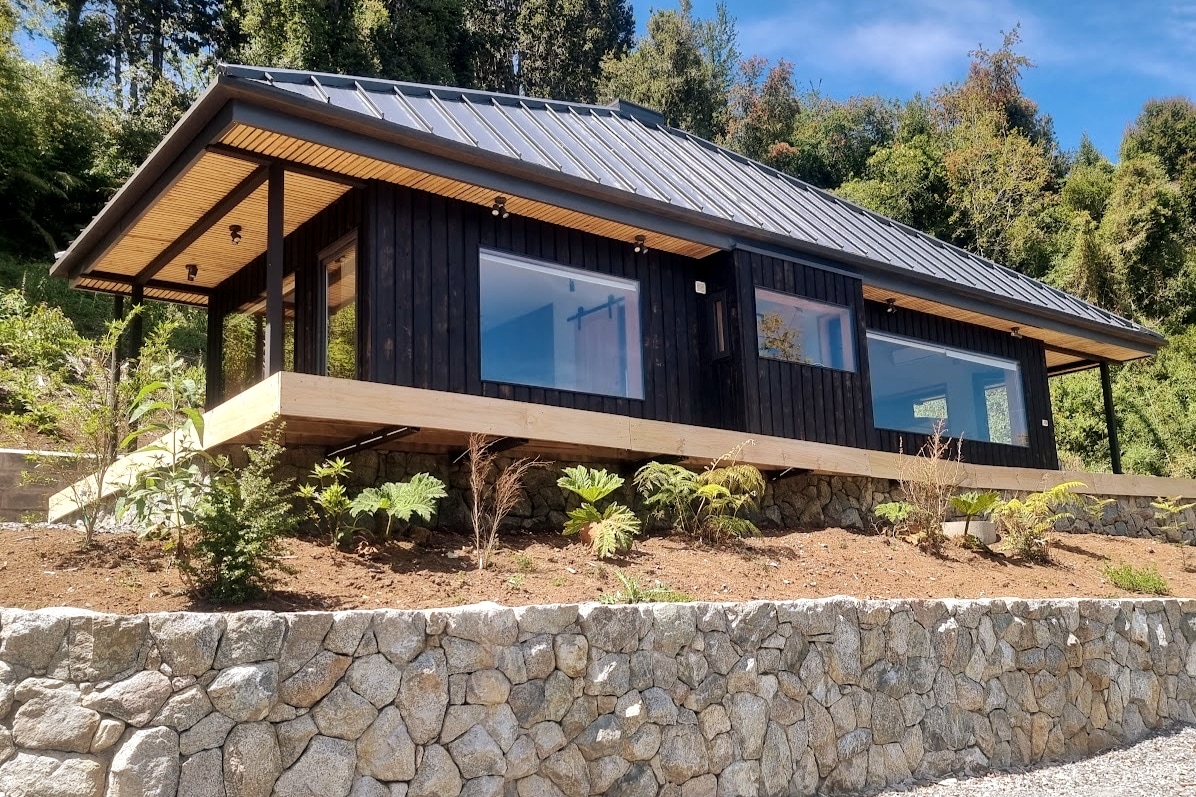 A modern black wooden house stands on a gentle slope, surrounded by native vegetation. Large windows provide ample natural light, while a wooden deck extends outwards, inviting outdoor relaxation. The structure is complemented by a stony foundation, enhancing the connection to the natural landscape.