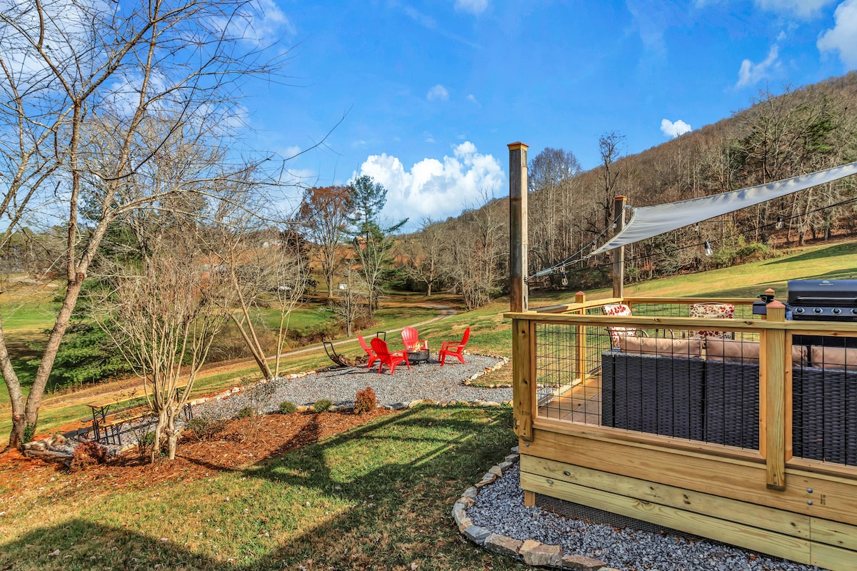 An outdoor deck area is shown, featuring a gravel pathway leading to red chairs arranged around a fire pit. A shaded seating arrangement is visible on one side, with a view of the surrounding grassy landscape and distant hills under a mostly clear sky.