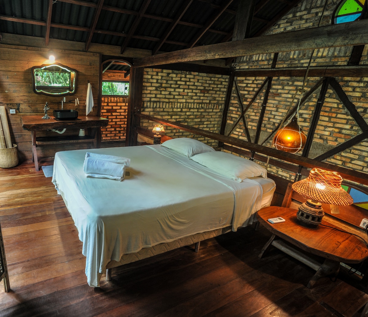 A spacious sleeping area features a large bed with crisp white linens and neatly arranged towels. Warm wooden floors complement the rustic wooden beams and brick walls. Soft lighting is provided by bedside lamps, and a small desk is visible in the corner.
