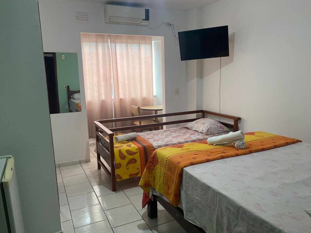 A spacious room accommodates a double bed and a single bed, both with fresh linens. Natural light filters through sheer curtains, enhancing the inviting ambiance. A wall-mounted television is visible, along with a small table and chair in the corner, providing a comfortable seating area.