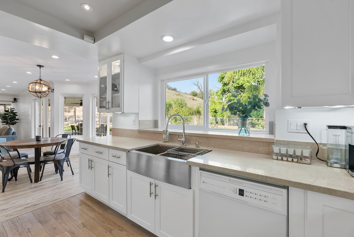 A modern kitchen features white cabinetry and a spacious countertop, illuminated by natural light from a large window. A double basin sink is situated in front of the window, providing a view of the landscape outside. A dining area is visible in the background.