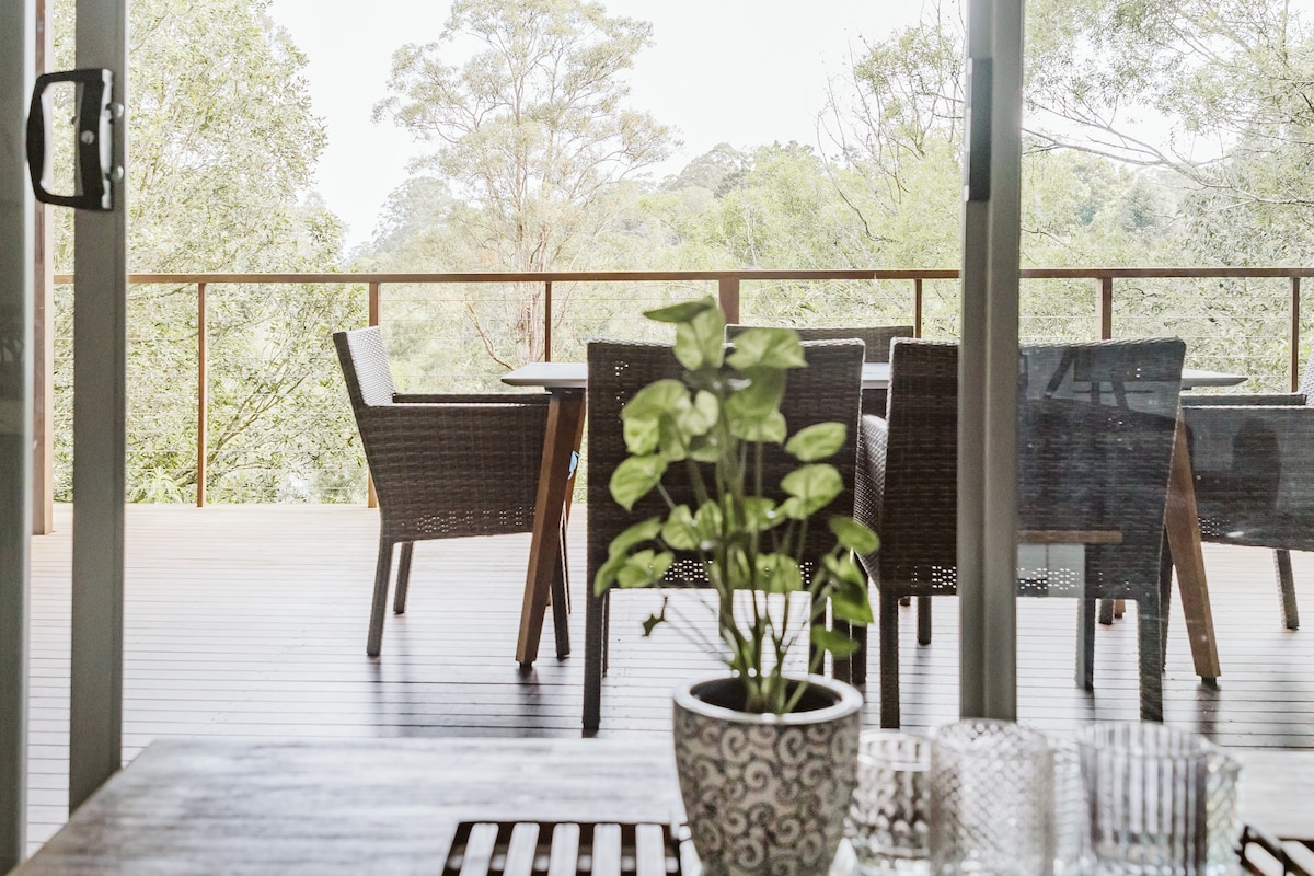 A spacious deck is visible through large glass doors, featuring a wooden dining table surrounded by wicker chairs. Lush greenery is present in the background, enhancing the connection to nature. A potted plant with broad leaves is positioned in the foreground.