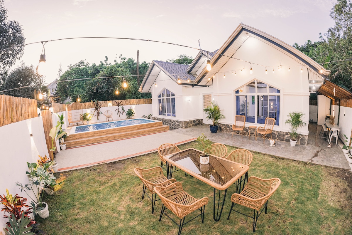 The outdoor space features a private pool surrounded by grass and ornamental plants. A dining table with several chairs is positioned nearby under string lights, enhancing the area for gatherings. The house showcases large windows, allowing natural light to fill the interior.