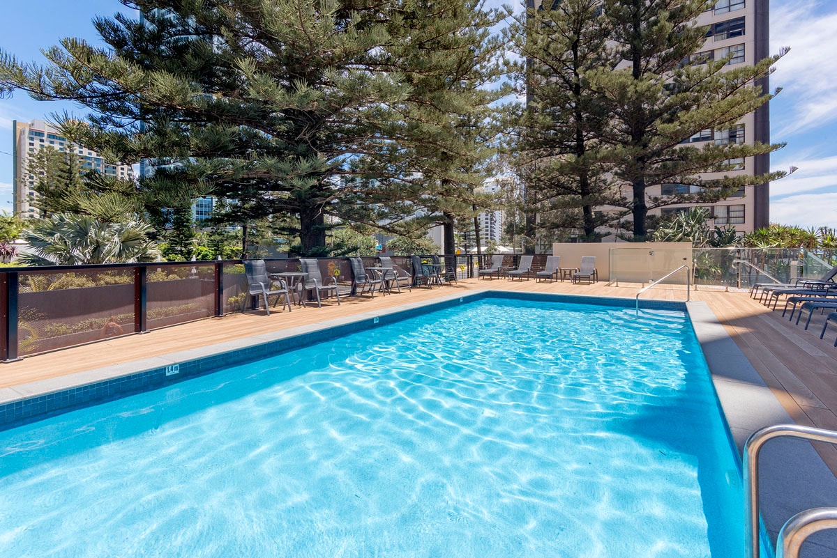 A swimming pool is surrounded by a wooden deck and lush greenery, with tall pine trees providing shade. Lounge chairs are positioned around the pool, allowing for relaxation. The clear blue water invites guests for a refreshing dip on a sunny day.