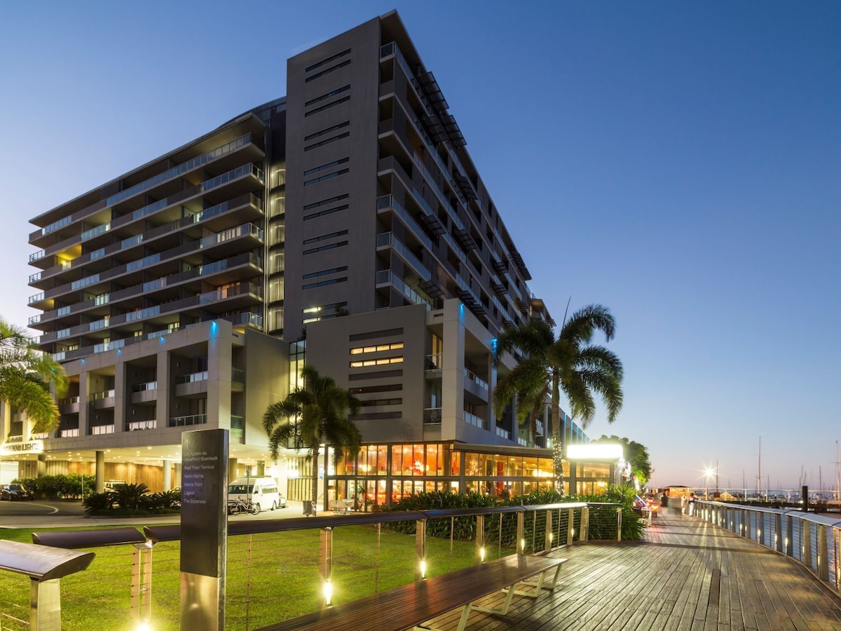 The modern apartment-style hotel is situated along the waterfront, featuring multiple levels and balconies that overlook the marina. A spacious wooden boardwalk runs adjacent to the building, bordered by greenery and palm trees, with soft lighting enhancing the evening ambiance.