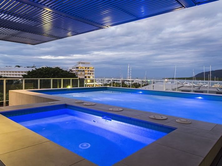A rooftop pool area is depicted, featuring two distinct pools with vibrant blue water. Soft evening lighting illuminates the space, creating a serene atmosphere. Views of the marina and boats beyond are visible, underscoring the waterfront location.