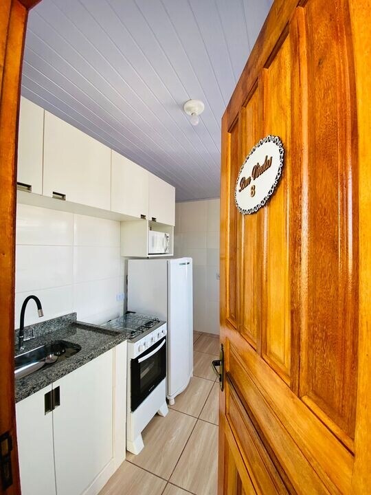 A modern kitchen space is visible through an open wooden door. It features sleek white cabinetry, a countertop with a sink, a microwave, and a refrigerator. A stove is positioned under a range hood, providing a functional cooking area.
