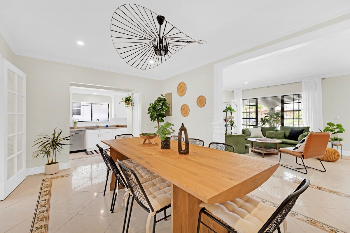 A modern dining area features a long wooden table surrounded by dining chairs. A large light fixture hangs above, and decorative wall elements add interest. Nearby, a spacious living area is visible, showcasing plush green seating and natural light filtering through large windows.