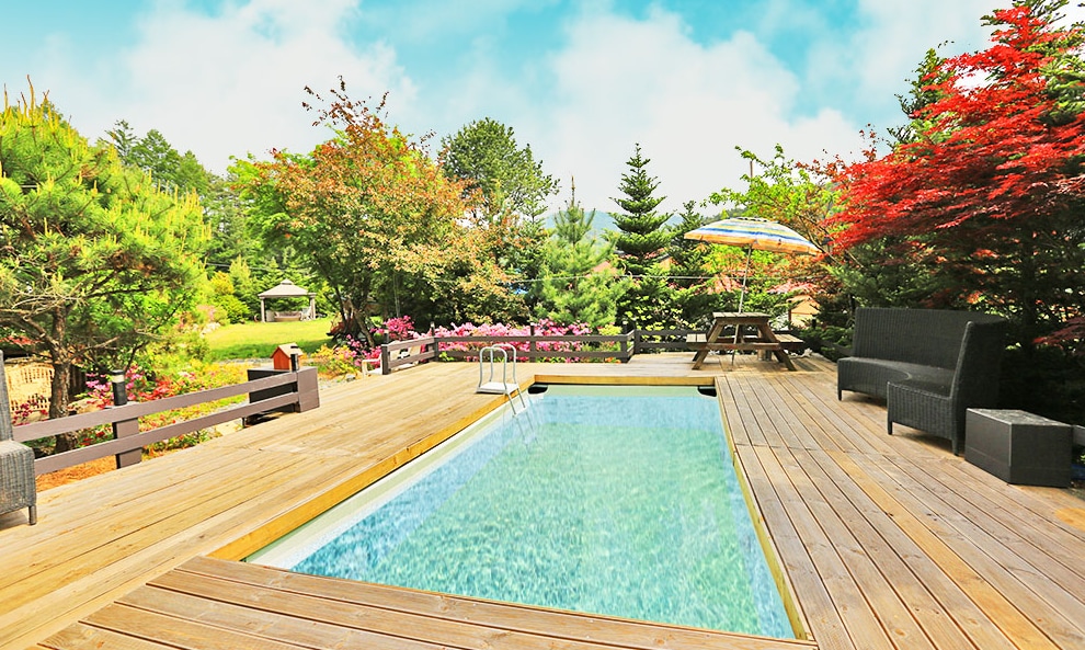 A serene outdoor area features a wooden deck surrounding a clear swimming pool. Lush greenery and colorful shrubs lend a vibrant backdrop, while comfortable seating areas are positioned nearby. An umbrella provides shade for relaxation, enhancing the inviting outdoor atmosphere.