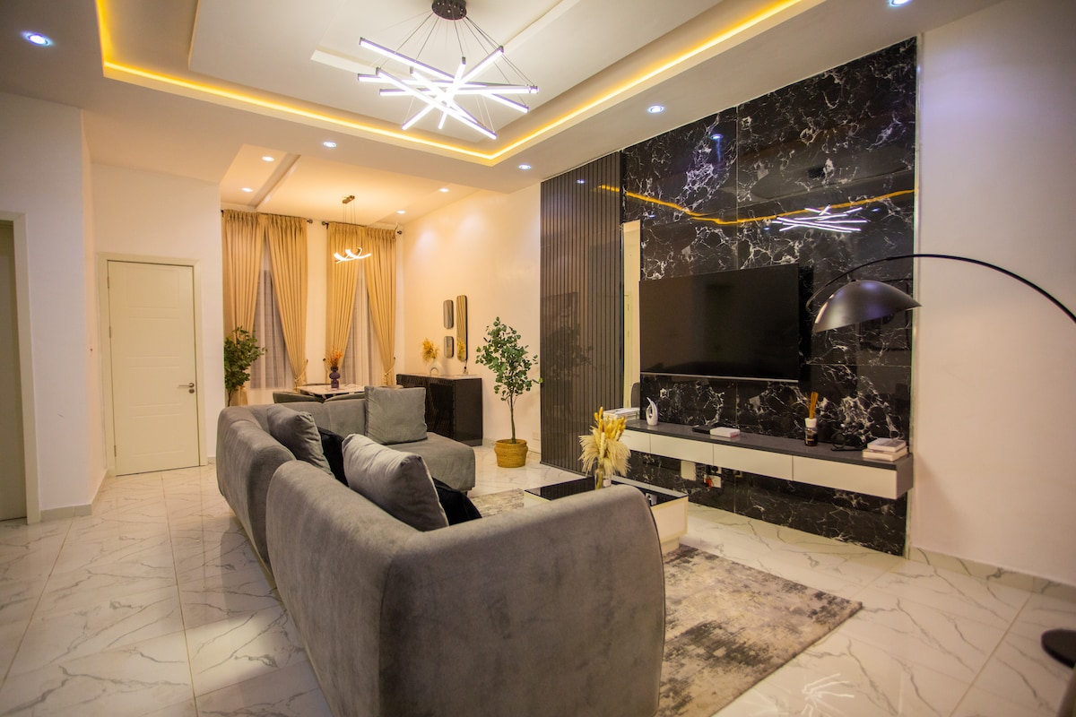 A contemporary living area features a plush gray sofa positioned facing a wall-mounted flat-screen TV. Soft lighting is provided by a modern ceiling fixture and floor lamp, enhancing the stylish decor. Neutral tones and textures create an inviting space, complemented by decorative plants.