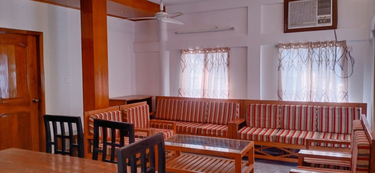 The living area features a set of striped couches arranged around a glass coffee table. Light streams through two large windows, framed by sheer curtains. A dining table with dark chairs complements the warm wooden tones of the space, enhanced by wooden ceiling beams.